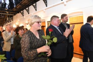 Publiczność bije brawa. Wszyscy stoją. Na pierwszym planie kobieta trzymająca w rękach różę