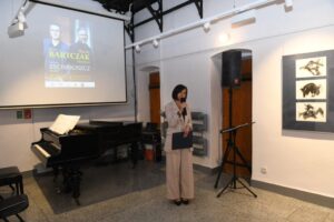 Kustosz muzeum PK, pani Lilianna Lewandowska z mikrofonem. Za nią stoi fortepian i znajduje się ekran, na który wyświetlony jest plakat wydarzenia