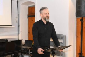 Maciej Bartczak stoi przed pulpitem. Ma czarną koszulę, siwiejące włosy i zarost