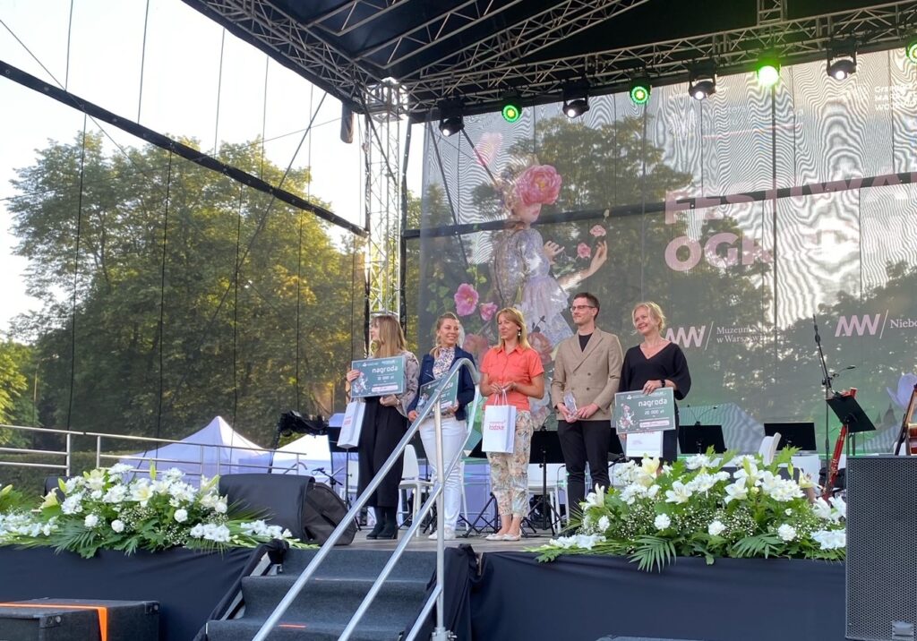 Grupa ludzi na scenie. Trzy osoby trzymają w rękach dyplomy, jedna statuetkę i jedna białą torbę papierową. Scena ozdobiona bukietami białych kwiatów.
