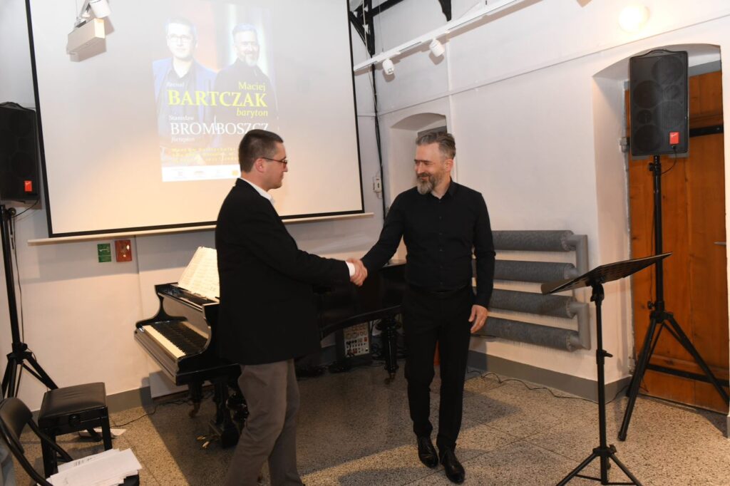 Dwóch elegancko ubranych mężczyzn (Maciej Bartczak i Stanisław Bromboszcz) podają sobie ręce. Za nimi fortepian i ekran, na którym wyświetla sie plakat wydarzenia
