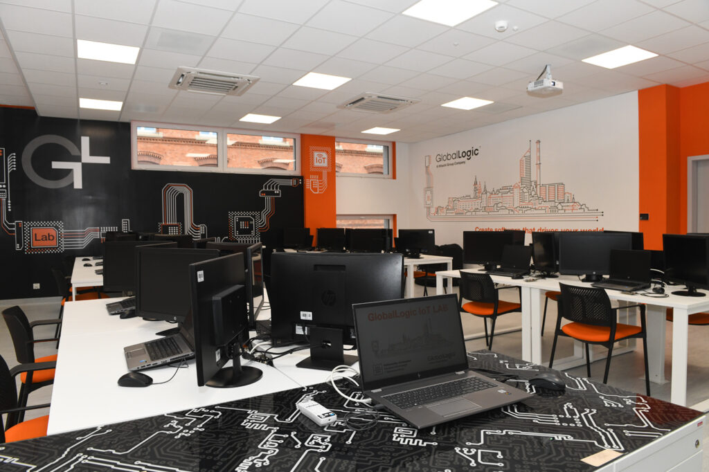 Sala dydaktyczna. Na stolikach rozłożone laptopy, monitory, myszki i klawiatury. Przy stolikach krzesła. W czarne, białe i pomarańczowe ściany. 