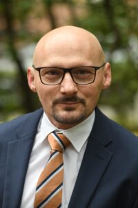 Dr Gancarzewicz patrzy w obiektyw, na twarzy lekki uśmiech, okulary. Ubrany w marynarkę, białą koszulę i krawat