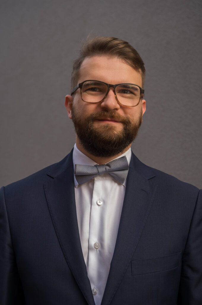 Mężczyzna około 40 lat w okularach i z zarostem, w marynarce, białej koszuli, pod muchą. Lekko uśmiecha się do zdjęcia. To dziekan Paweł Pławiak