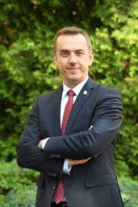 Rektor Andrzej Szarata na zdjęciu portretowym. Ubrany w garnitur, ręce splecione na piersiach, patrzy pewnym wzrokiem w obiektyw