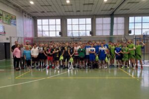 Wspólne zdjęcie zawodników wszystkich drużyn biorących udział w zawodach. Wszyscy chłopcy mają sportowe stroje. Wraz z nimi pozują do zdjęcia dwie kobiety i dwóch mężczyzn. Zdajecie zrobione na sali gimnastycznej