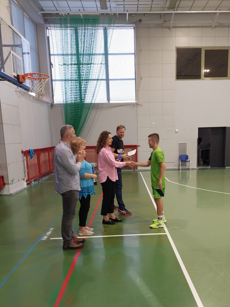 Sala gimnastyczna. Kobieta w różowej bluzce i czarnych spodniach wręcza coś chłopakowi w sportowym stroju. Obok niej stoi dwóch mężczyzn i kobieta