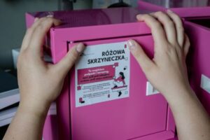 Widoczne dłonie, które naklejają na różową skrzynkę naklejkę informacyjną