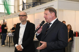 Dwóch mężczyzn w garniturach – prof. Bogdał i przedstawiciel ABB. Prorektor ds. nauki przemawia do mikrofonu. W tle stoiska i pojedynczy ludzie