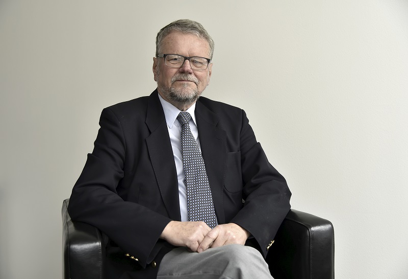 Czeski profesor pozuje do zdjęcia na siedząco. Siwa, krótka broda, okulary. Elegancko ubrany, czarna marynarka, krawat.