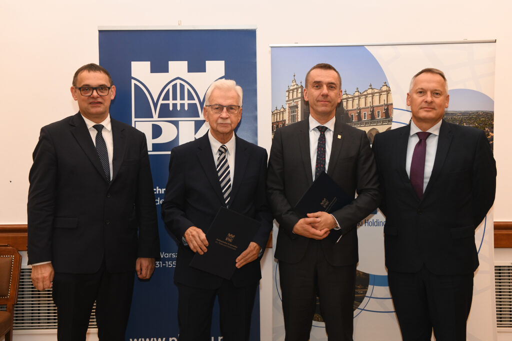 Czterech mężczyzn pozuje do zdjęcia na tle rolupów Politechniki Krakowskiej i Krakowskiego Holdingu Komunalnego