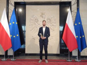 Łukasz Ślaga ubrany w garnitur pozuje do zdjęcia ze statuetką w rękach, w tle duży orzeł RP, a po prawej i lewej stronie laureata flagi Polski i UE