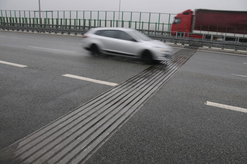 Biały samochód pędzi po asfaltowej drodze. Jest lekko rozmazany, co sugeruje dużą prędkość z jaką jedzie.