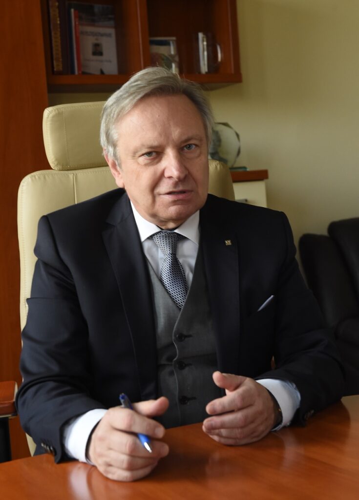 Jerzy Sładek siedzi  przy biurku, patrzy w obiektyw, ręce na blacie, między palcami długopis