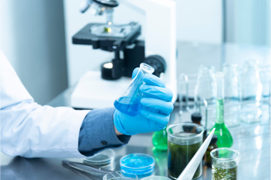 Ręce w fartuchu, rękawiczki lateksowe na dłoniach, probówka w palcach. Dookoła na stoliku naczynia laboratoryjne, w tle mikroskop