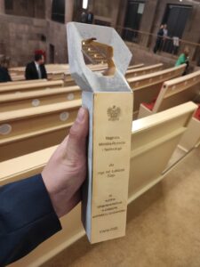 Na zdjęciu statuetka trzymana w ręce. Statuetka to bryłka z wygrawerowanym napisem: Nagroda Ministra Rozwoju i Technologii dla mgr. inż. Łukasza Ślagi za wybitne osiągnięcia twórcze w dziedzinie architektury i budownictwa