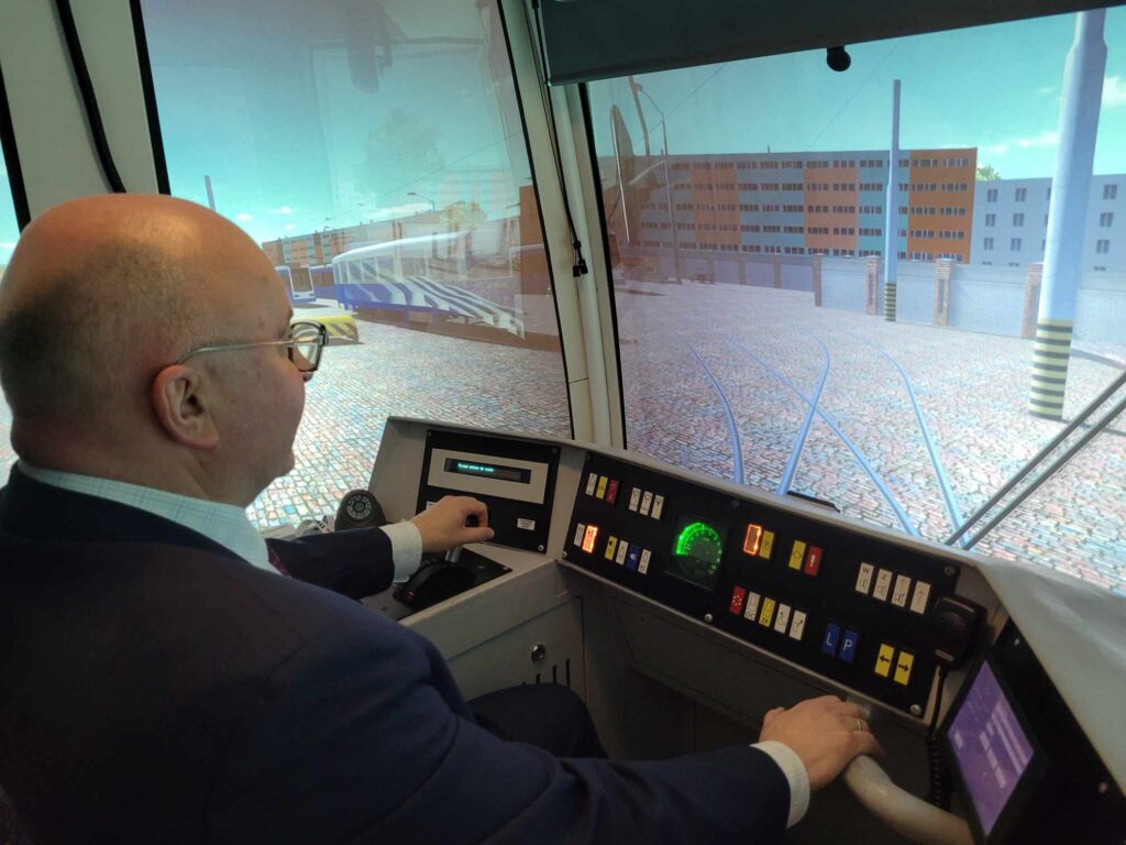 Mężczyzna przy pulpicie symulującym pulpit sterowniczy tramwaju, przed nim projekcja przedstawiająca trasę tramwajową