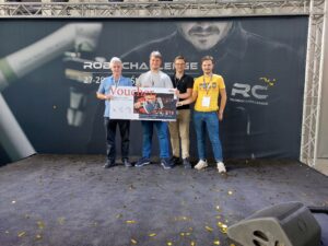 Czterech mężczyzn. Jeden w średnim wieku z siwymi włosami i trzech studentów. W rękach trzymają duży voucher