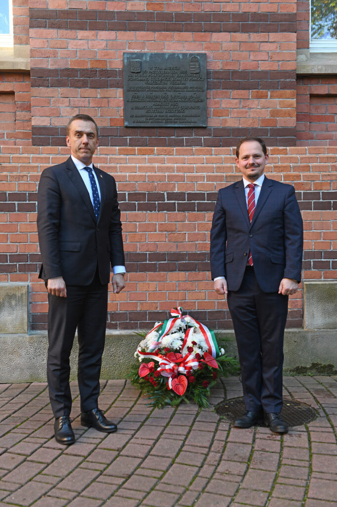 Rektor PK prof. Andrzej Szarata i konsul generalny Węgier w Krakowie dr Tibor Gerencsér pozują do zdjęcia, w tle tablica na ścianie budynku, która upamiętnia wydarzenia z 1956 r., na dole kwiaty złożone przez konsula i rektora