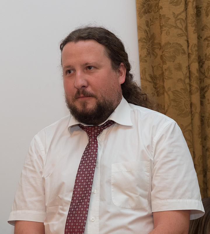 Mężczyzna w średnim wieku z jasną brodą i długimi, kędzierzawymi włosami związanymi w kucyk. Ubrany jest w jasną koszulę z krótkim rękawem. Ma czerwony krawat. To doktor Adam Gska