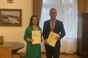Kobieta w zielonej sukience i mężczyzna w garniturze pozują do zdjęcia. W dłoniach trzymają podpisane umowy