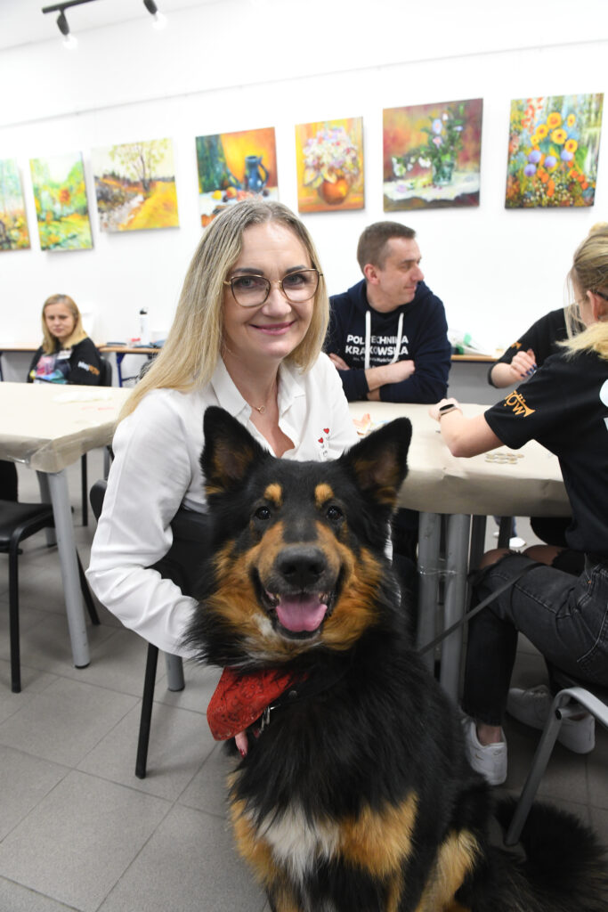 Blondynka w okularach i białej koszuli przytula dużego psa w typie owczarka niemieckiego. W tle widać wolontariuszy i rektora Szaratę