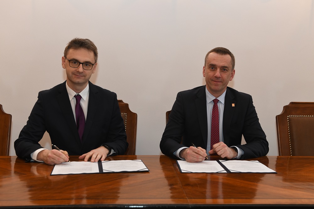 Marcin Karp i prof. Andrzej Szarata pozują do zdjęcia. Siedzą przy stole, trzymają w rękach długopisy. Na stole leżą umowy