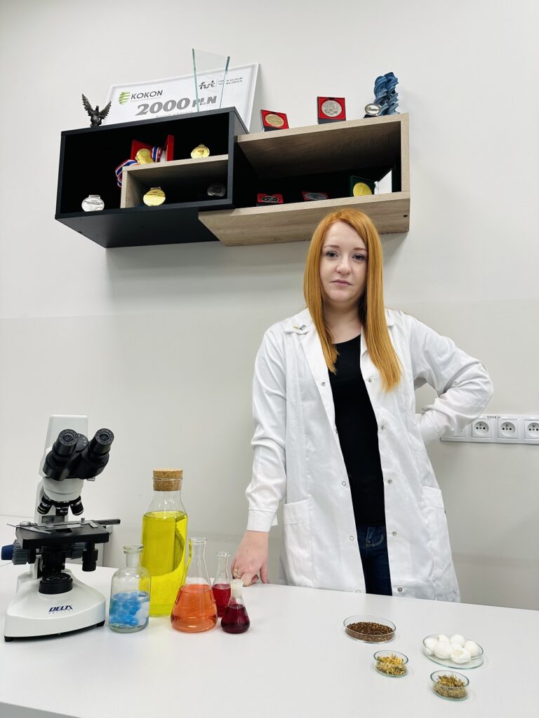 Magdalena Bańkosz w kiltu chemika pozuje do zdjęcia. Na stole przed nią mikroskop i naczynia z kolorowymi płynami.  Za nią, na półce ściennej m.in. medale i dyplomy za osiągnięcia naukowe