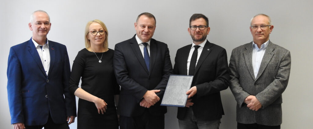Grupa osób pozuje do wspólnego zdjęcia. Dziekan Beńko trzyma egzemplarz umowy w skórzanej teczce