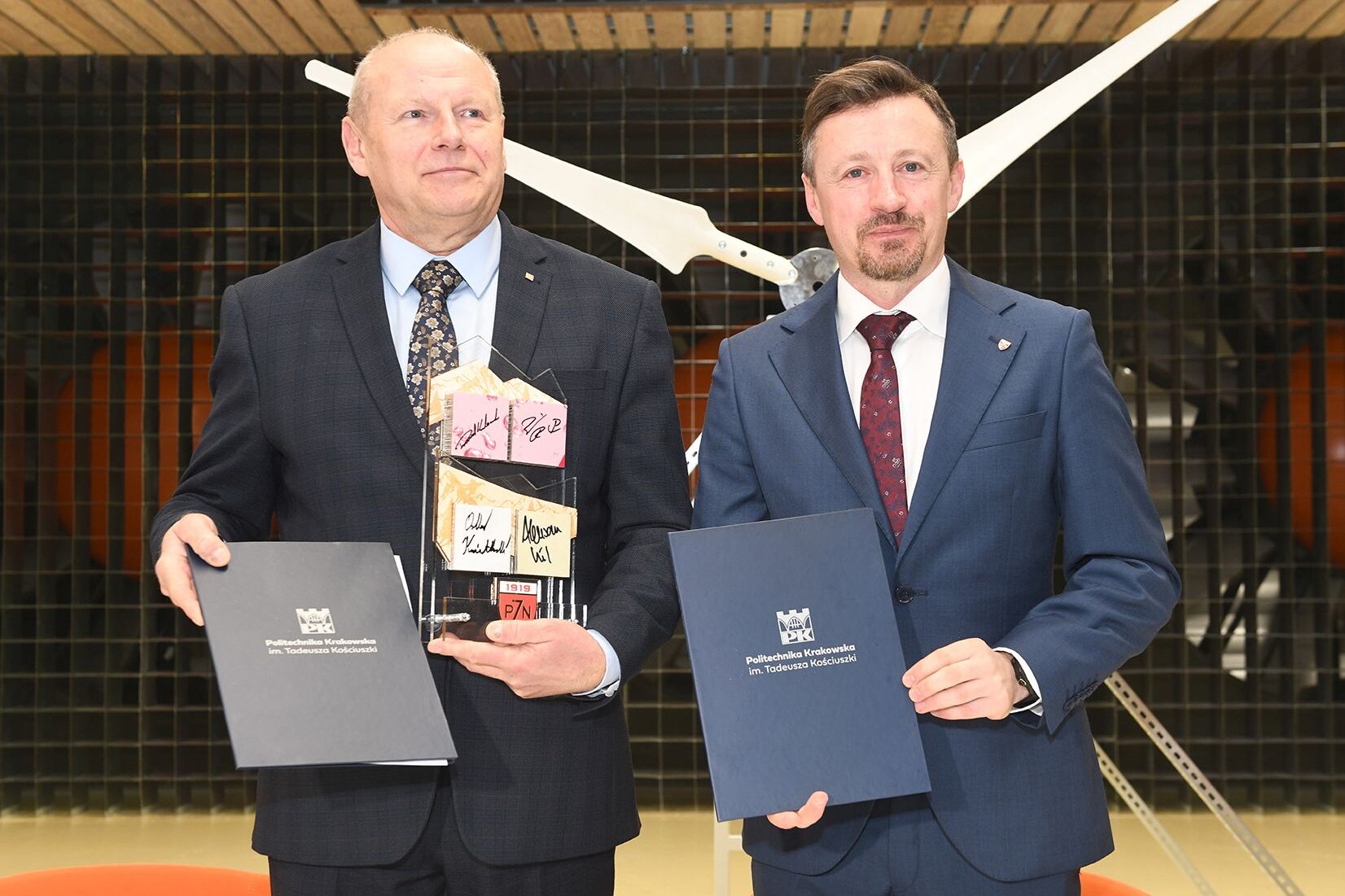 Tomasz Kapecki i Adam Małysz pozują do zdjęcia trzymając w ręce egzemplarze umów. Prorektor pokazuje również prezent od PZN - konstrukcję na drewnie z autografami skoczków i innych sportowców